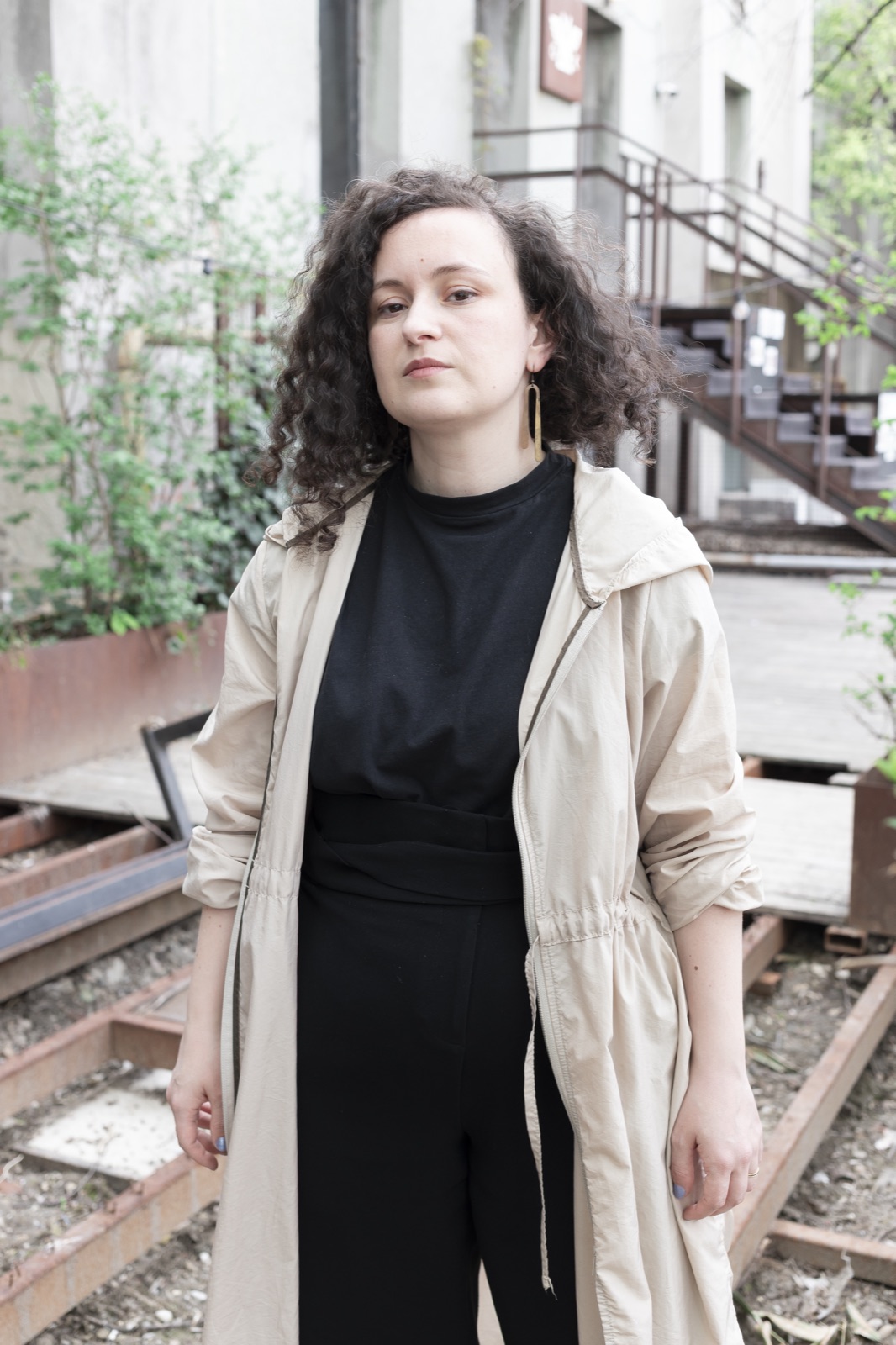 Elena Zara standing in a courtyard in a pale coat, looking at the camera.