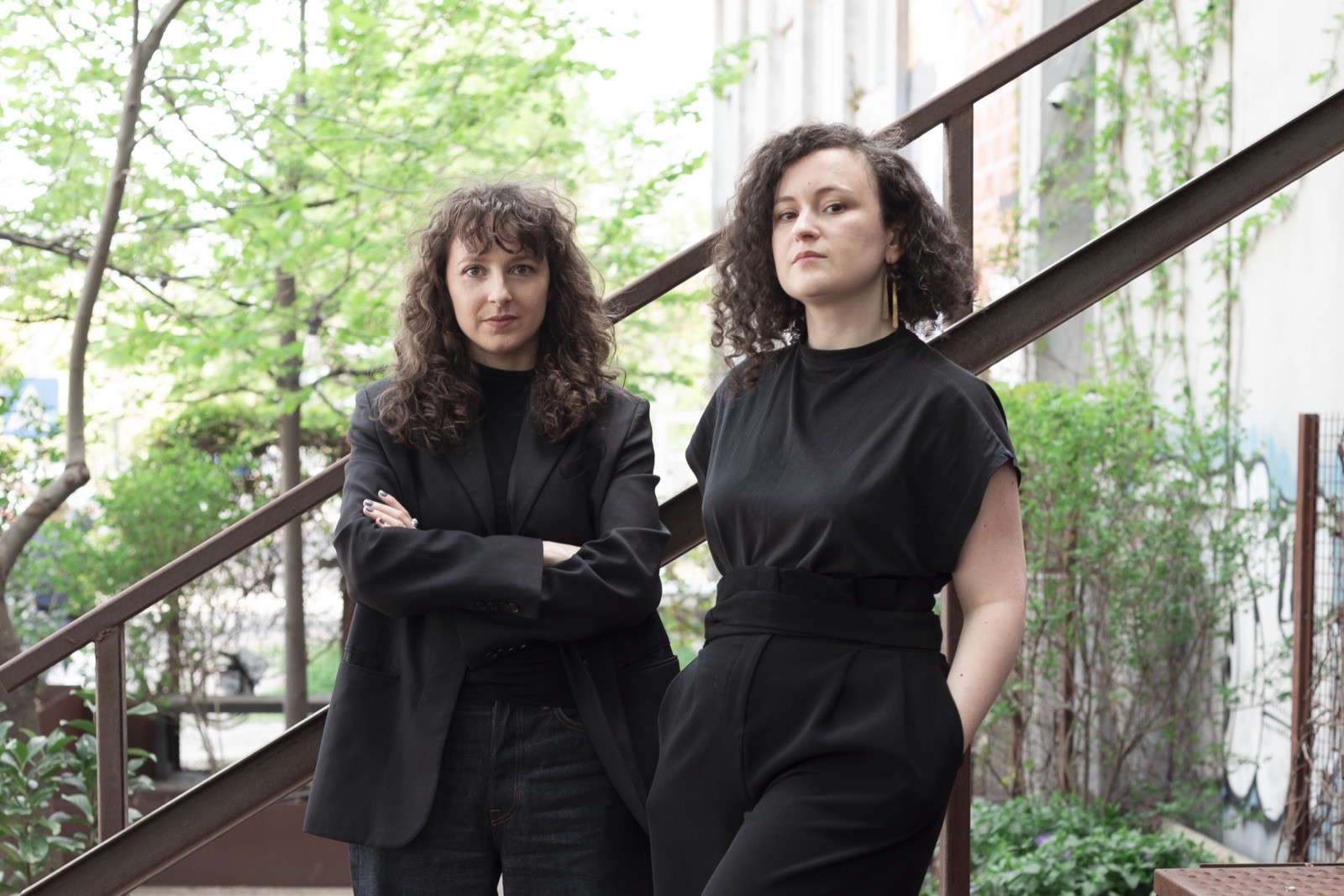 Andra Ionel and Elena Zara, the two founders of ZEIA studio, standing together by a rusted steel staircase in a green courtyard.